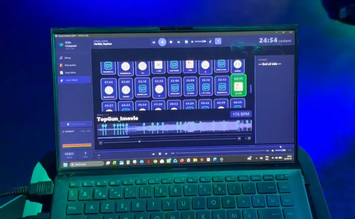 A laptop at the front of an indoor cycling studio running RideComposer Coach Mode, showing the grid of cues, the track playing and the time remaining